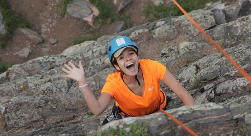 From above, a person wearing safety gear smiles and waves as they climb towards the camera. 